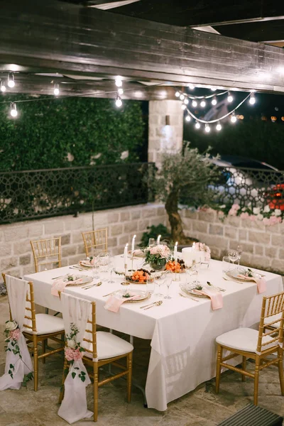 Δεξίωση τραπεζιού γάμου. Τραπέζι ορθογώνιο για έξι άτομα με λευκό τραπεζομάντηλο και ροζ χαρτοπετσέτες. Το χρυσό chairofchia είναι διακοσμημένο με ύφασμα και ροζ λουλούδια. Η γιρλάντα καίει πάνω από τραπέζι. — Φωτογραφία Αρχείου