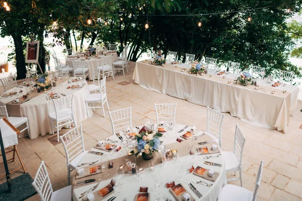 Bruiloft diner tafel receptie. Ronde tafels met witte tafelkleden, bruine lopers, boeketten bloemen, Chiavari stoelen, menu zonde-ingesloten in servet. Restaurant buiten, in het bos, waterverlichting bloemenslingers — Stockfoto