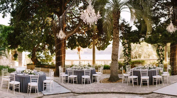 Düğün masası resepsiyonu. Gri masa örtüsü olan misafirler için yuvarlak ziyafet masaları. Çiçek aranjmanı masanın ortasına. Ağaç gövdelerinde çelenkler, havada kristal avizeler