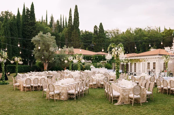 Recepción de mesa de cena de boda. Elegantes mesas para invitados con manteles color crema con estampados, sobre césped verde, con guirnaldas y candelabros colgando sobre ellas. Sillas con respaldo redondo — Foto de Stock