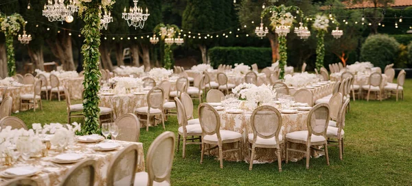 結婚式のディナーテーブルレセプション。緑の芝生の上に、ガーランドやシャンデリアがかかっているパターンのクリームテーブルクロスを持つお客様のためのエレガントなテーブル。丸みを帯びた椅子 — ストック写真