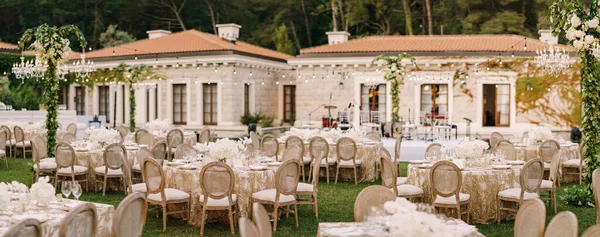 Hochzeitstisch-Empfang. Elegante Tische für Gäste mit cremefarbenen Tischdecken mit Mustern auf grünem Rasen, über denen Girlanden und Kronleuchter hängen. Stühle mit rundem Rücken — Stockfoto