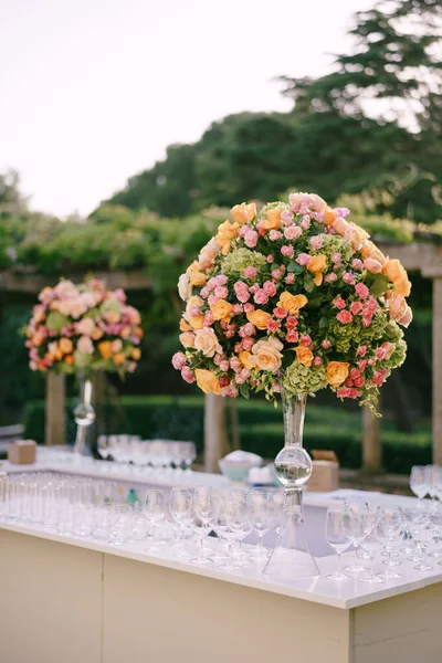 Recepción de mesa de cena de boda. Enormes ramos de rosas amarillas, rosadas, naranjas y hortensias verdes en un jarrón de cristal en una pierna alta. Ramos grandes en forma de bola. Un bar con muchos vasos . — Foto de Stock