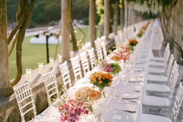 Hochzeitstisch-Empfang. Ein sehr langer Hochzeitstisch, der ins rechte Licht gerückt wird. Sträuße von gelben, orangen und rosa Rosen auf Tischen. Glasstühle Chiavari, Tiffany aus transparentem Kunststoff — Stockfoto