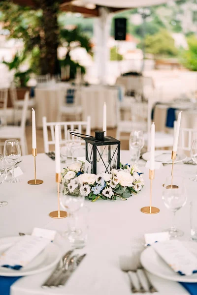 Recepción de mesa de cena de boda. Un candelabro de metal en el centro de la mesa rodeado de composición floral, velas en candelabros de oro, mesas blancas y platos con servilletas azules — Foto de Stock