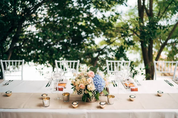 Resepsi meja makan malam pernikahan. Sebuah karangan bunga mawar merah muda dan hidrangea biru ada di atas meja dalam vas. Pelari krem dengan lilin yang menyala. Empat kursi Chiavari di meja persegi panjang. — Stok Foto
