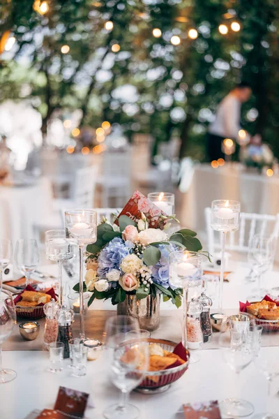 Wedding dinner table reception. Bouquet in a vase on a table of pink and white roses and blue hydrangea, with the number of the table on the plate. Burning candles in high glass candlesticks. — Stock Photo, Image