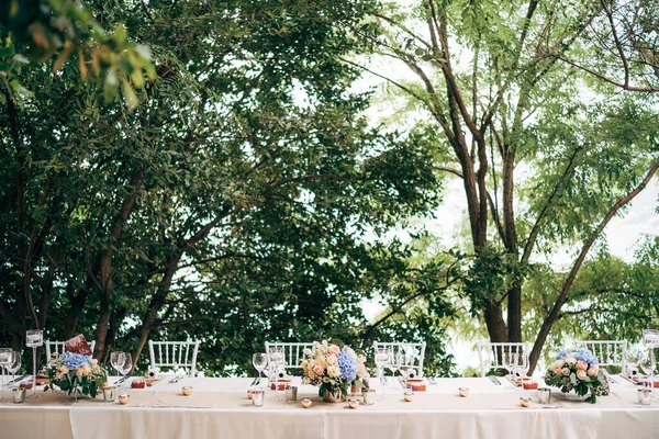 Recepción de mesa de cena de boda. Un ramo de rosas rosadas y hortensias azules está sobre la mesa en un jarrón. Corredor beige con velas encendidas. Seis sillas Chiavari en una mesa rectangular . —  Fotos de Stock