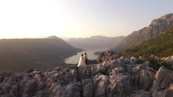 Nevěsta a ženich stojí na vrcholu hory Lovchen v Černé Hoře s fantastickým výhledem na Kotor Bay, Kotor staré město, Prcanj, Muo, na vrcholky hor a lesů, zaplaven západem slunce — Stock video