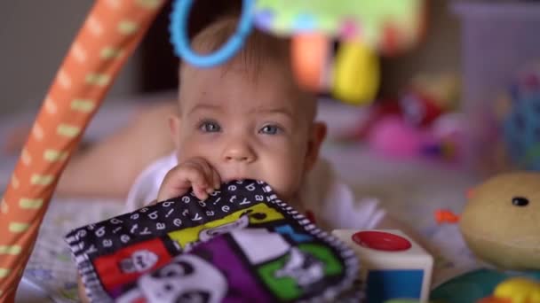 Bebé niña se acuesta sobre su estómago y mordisquea en un libro de desarrollo en una estera de desarrollo, mirando a la cámara — Vídeo de stock