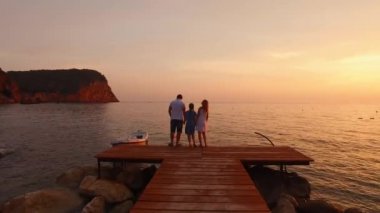 Bir aile ahşap bir deniz iskelesinde el ele tutuşmuş, denizin üzerindeki renkli, yakıcı günbatımını seyrediyor. Aile değerleri, sevgi ve birlik kavramı. Karadağ 'da aile tatilleri Adriyatik' te.