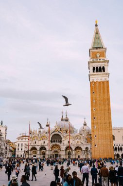 Piazza San Marco 'daki kırmızı tuğladan yapılmış büyük bir çan kulesi Venedik, İtalya' daki St. Marks Katedrali 'nin Campanile' i. İki martı uçuyor. Meydandaki kulenin eteklerinde turist kalabalığı var..