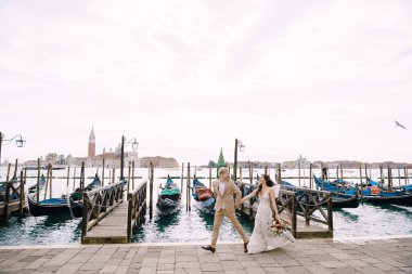 Gelin ve damat, San Giorgio Maggiore 'a ve günbatımı gökyüzüne tepeden bakan Venedik' teki Piazza San Marco yakınlarındaki gondol iskelesinde el ele koşuyorlar. Venedik, İtalya 'daki en büyük gondol iskelesi..