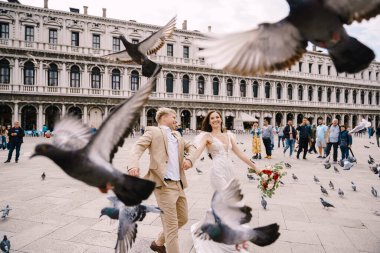 Venedik Düğünü, İtalya. Gelin ve damat Piazza San Marco 'da, turist kalabalığıyla çevrili Ulusal Arkeoloji Müzesi Venedik' in ortasında uçan güvercin sürüsünün arasında koşuyorlar..