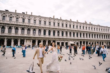 Venedik Düğünü, İtalya. Gelin ve damat Piazza San Marco 'da, turist kalabalığıyla çevrili Ulusal Arkeoloji Müzesi Venedik' in ortasında uçan güvercin sürüsünün arasında koşuyorlar..