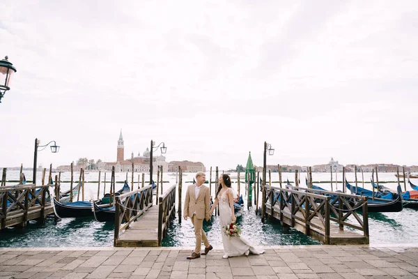 A menyasszony és a vőlegény sétálnak végig a gondola mólón, kéz a kézben Velencében, közel a Piazza San Marco, kilátással a San Giorgio Maggiore és a naplemente ég. Velence legnagyobb gondola mólója, Olaszország. — Stock Fotó