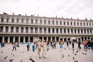 Venedik Düğünü, İtalya. Gelin ve damat Piazza San Marco 'da, turist kalabalığıyla çevrili Ulusal Arkeoloji Müzesi Venedik' in ortasında uçan güvercin sürüsünün arasında koşuyorlar..