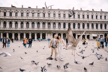 Venedik Düğünü, İtalya. Gelin ve damat Piazza San Marco 'da, turist kalabalığıyla çevrili Ulusal Arkeoloji Müzesi Venedik' in ortasında uçan güvercin sürüsünün arasında koşuyorlar..