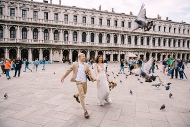 Venedik Düğünü, İtalya. Gelin ve damat Piazza San Marco 'da, turist kalabalığıyla çevrili Ulusal Arkeoloji Müzesi Venedik' in ortasında uçan güvercin sürüsünün arasında koşuyorlar..
