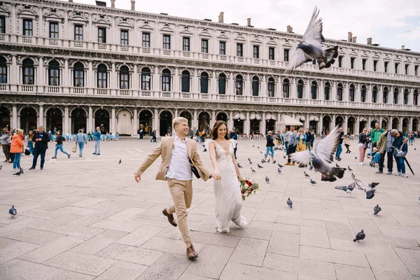 Velencei esküvő, Olaszország. A menyasszony és a vőlegény egy csapat repülő galambon futnak keresztül a Piazza San Marco-n, a Velencei Nemzeti Régészeti Múzeumban, körülvéve turisták tömegével.. — Stock Fotó