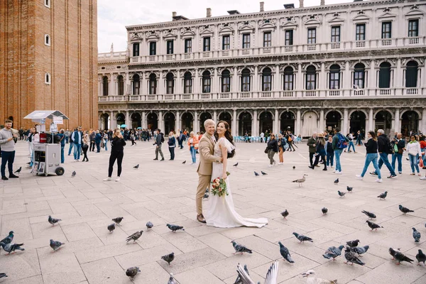 Wedding in Venice, Italy. Bride and groom are looking at camera among the pigeons in Piazza San Marco, against backdrop of the National Archaeological Museum Venice, surrounded by a crowd of tourists. — Stock Photo, Image