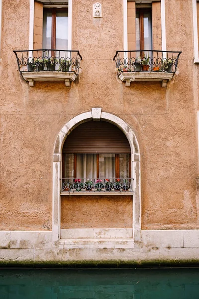 Будівництво фасадів у Венеції, Італія. Кам'яна арка над дерев'яним вікном на фасаді будівлі. Білі завіси у вікні, маленькі квіти у горщиках на підвіконні з металевим ковані парканом. — стокове фото
