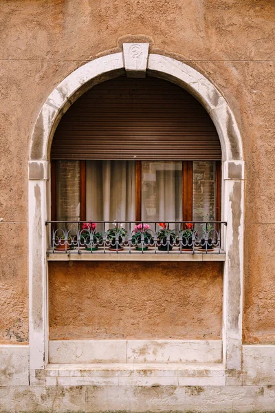 Close-ups de fachadas de edifícios em Veneza, Itália. Um arco de pedra acima de uma janela de madeira na fachada do edifício. Cortinas brancas na janela, pequenas flores em vasos no peitoril da janela com uma cerca forjada de metal — Fotografia de Stock