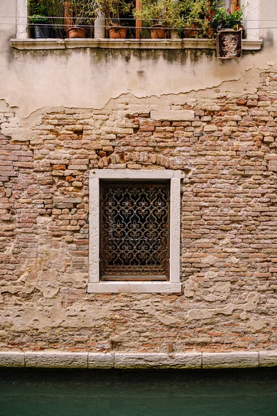 Будівництво фасадів у Венеції, Італія. Цегляна кам'яна стіна з вікном, повністю закрита металевою ковані решіткою, в невеликому каналі між будинками. Пальці з квітами стоять на підвіконні. — стокове фото