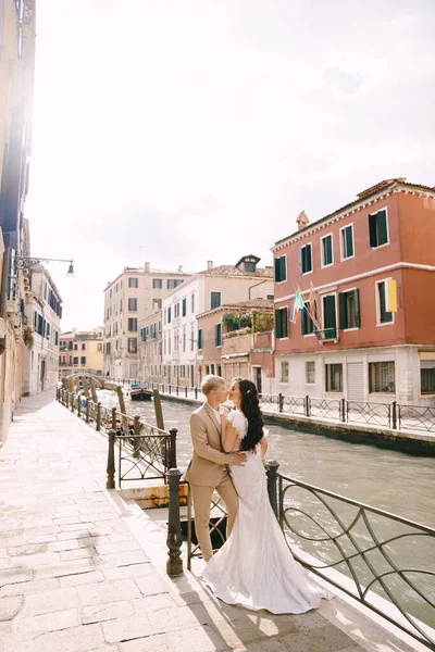 Italië bruiloft in Venetië. Pasgetrouwden staan aan de oevers van het kanaal van Venetië. De bruidegom knuffelt de bruid bij de taille. Witte trouwjurk met kleine mooie trein en zandkleurige heren pak. — Stockfoto