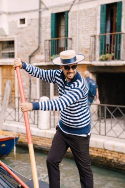 Venedik, İtalya - 04 Ekim 2019: Gülümseyen mutlu bir gondolcu, kürek çeken turistlerle dolu bir gondolu kontrol ediyor. Klasik mavi-beyaz çizgili ceket ve şapka tarihi bir görüntüdür..