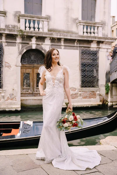 Velencei esküvő Olaszországban. Egy fehér ruhás menyasszony, egy vonattal, fehér és vörös rózsacsokorral a kezében, a mólón áll egy keskeny velencei csatornában, a kikötött gondola mellett.. — Stock Fotó
