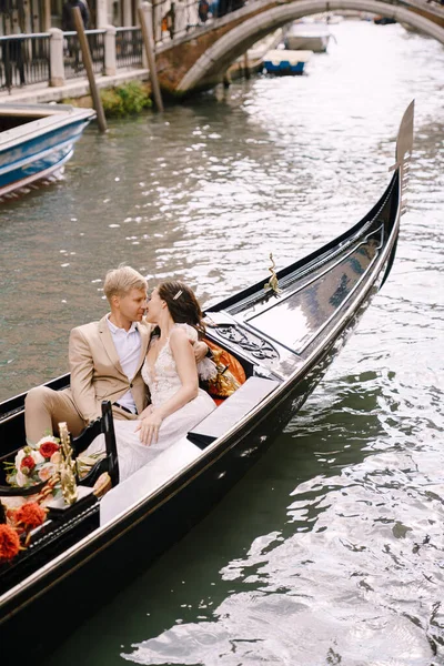 Italy wedding in Venice. The bride and groom ride in a classic wooden gondola along a narrow Venetian canal. Close-up of cuddles newlyweds. — Stock Photo, Image