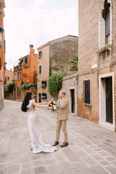 Olaszország esküvője Velencében. A menyasszony és a vőlegény a város elhagyatott utcáin sétálnak. A friss házasok ölelkeznek, táncolnak, fogják egymás kezét a festői vörös téglaházak hátterében.. — Stock Fotó