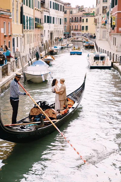 Italia boda en Venecia. Un gondolero enrolla a los novios en una clásica góndola de madera a lo largo de un estrecho canal veneciano. Recién casados están de pie en el barco, vista trasera . — Foto de Stock