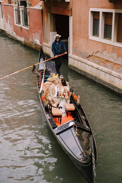 Italia boda en Venecia. Un gondolero enrolla a los novios en una clásica góndola de madera a lo largo de un estrecho canal veneciano. Los recién casados están sentados en un bote y quieren besarse. . — Foto de Stock