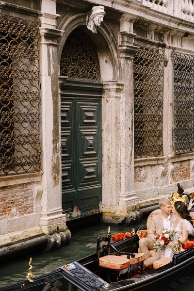 Mariage italien à Venise. Gondolier roule mariée et marié dans la gondole en bois classique le long du canal vénitien étroit. Les jeunes mariés sont assis dans le bateau nez à nez, nager sur fond de vieux treillis forgé — Photo