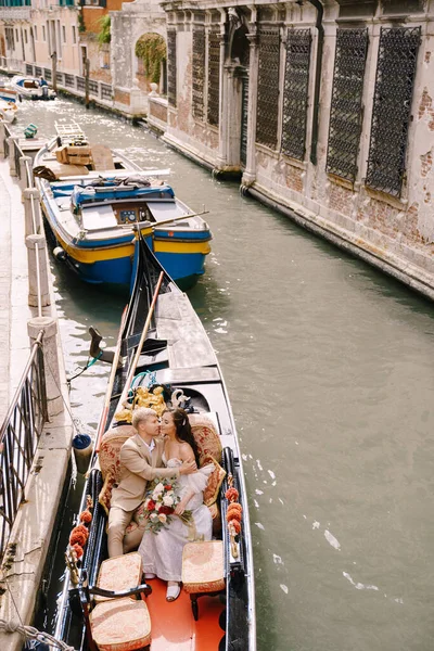 Italia boda en Venecia. Un gondolero enrolla a los novios en una clásica góndola de madera a lo largo de un estrecho canal veneciano. Recién casados se besan en un bote amarrado a la orilla . — Foto de Stock