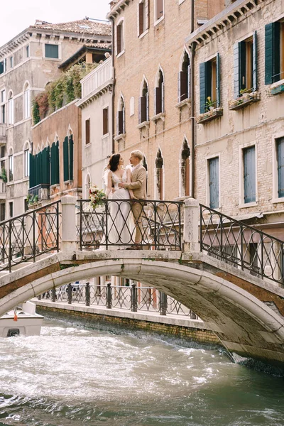 Italia boda en Venecia. La novia y el novio están de pie en un puente de piedra sobre un estrecho canal veneciano. Recién casados caminan por la ciudad tomando fotos en la calle . — Foto de Stock