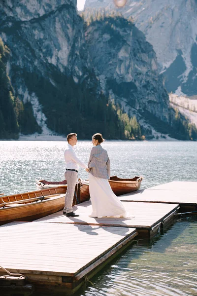 A menyasszony és a vőlegény sétálnak végig egy fa hajó dokk a Lago di Braies Olaszországban. Esküvő Európában, a Braies tónál. Friss házasok séta, csók, ölelés hátterében sziklás hegyek. — Stock Fotó