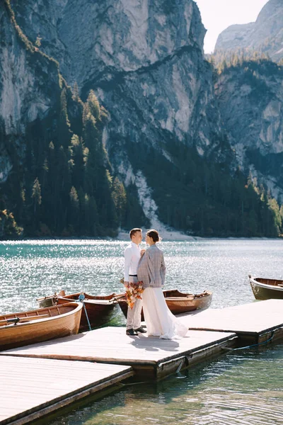 De bruid en bruidegom lopen langs een houten aanlegsteiger bij het Lago di Braies in Italië. Bruiloft in Europa, aan het Braies meer. Pasgetrouwden lopen, kussen, knuffelen op een achtergrond van rotsachtige bergen. — Stockfoto