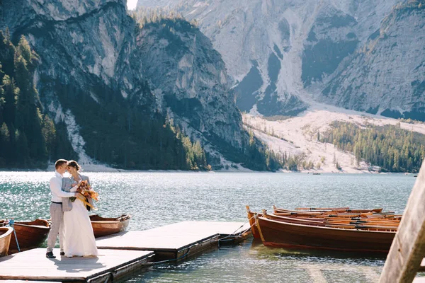Das Brautpaar spaziert an einem hölzernen Bootsanleger am Pragser See in Italien entlang. Hochzeit in Europa, am Pragser See. Frischvermählte spazieren, küssen, umarmen sich vor dem Hintergrund felsiger Berge. — Stockfoto