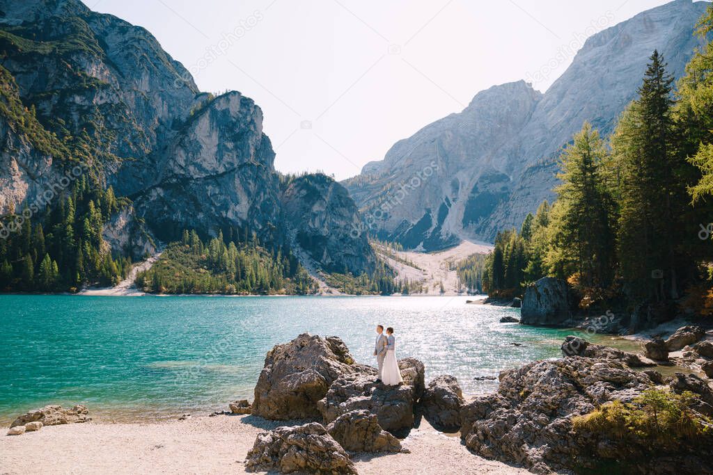 The bride and groom are standing on stones overlooking the Lago di Braies in Italy. Destination wedding in Europe, on Braies lake. Loving newlyweds walk against the backdrop of amazing nature.