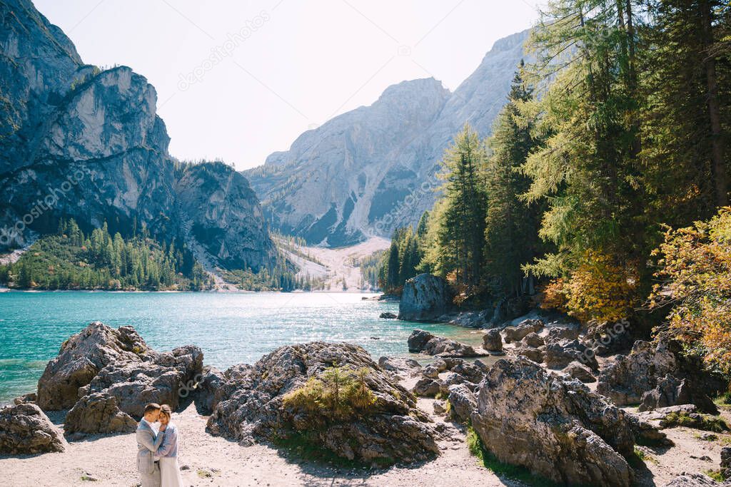 Bride and groom stand against the backdrop of stones overlooking Lago di Braies in Italy. Destination wedding in Europe, on Braies lake. Loving newlyweds walk against the backdrop of amazing nature.