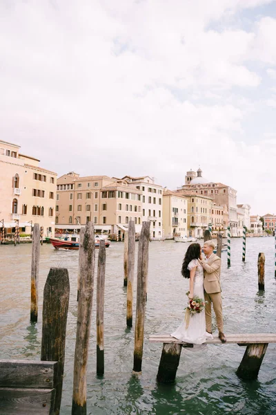 Mariage italien à Venise. Les mariés se tiennent debout sur une jetée en bois pour bateaux et gondoles, près des mâts d'amarrage rayés verts et blancs, sur fond de façades des bâtiments du Grand Canal . — Photo