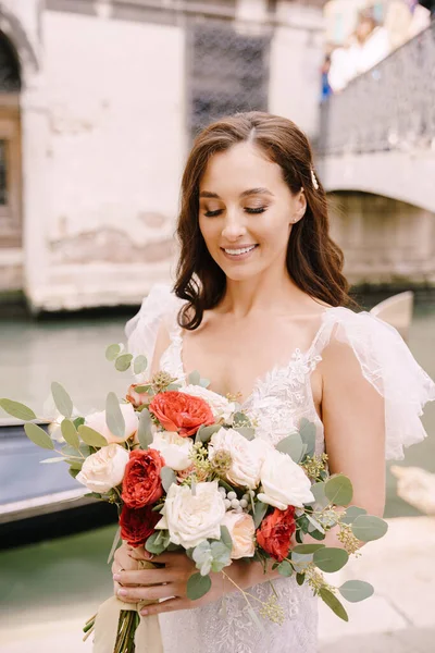 Venedigs bröllop i Italien. En brud i en vit klänning, med ett tåg, med en bukett vita och röda rosor i händerna, står på piren nära förtöjd gondol i en smal venetiansk kanal. — Stockfoto