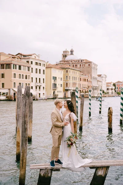 Olaszország esküvője Velencében. A menyasszony és a vőlegény áll egy fa mólón hajók és gondolák, közel a csíkos zöld és fehér kikötői oszlopok, háttér homlokzatok Grand Canal épületek. — Stock Fotó