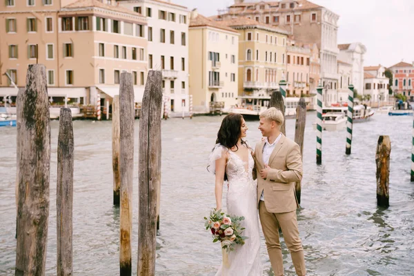 Olaszország esküvője Velencében. A menyasszony és a vőlegény áll egy fa mólón hajók és gondolák, közel a csíkos zöld és fehér kikötői oszlopok, háttér homlokzatok Grand Canal épületek. — Stock Fotó