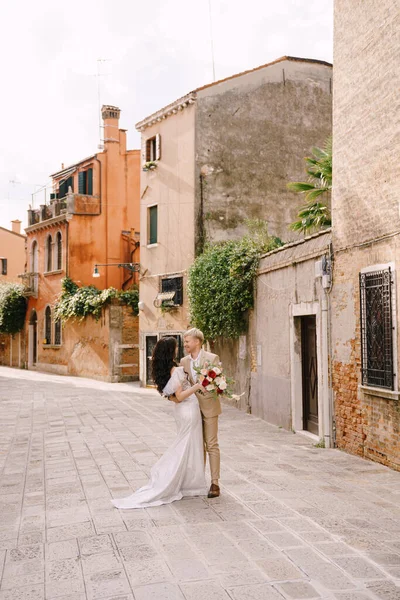 Olaszország esküvője Velencében. A menyasszony és a vőlegény a város elhagyatott utcáin sétálnak. A friss házasok ölelkeznek, táncolnak, fogják egymás kezét a festői vörös téglaházak hátterében.. — Stock Fotó