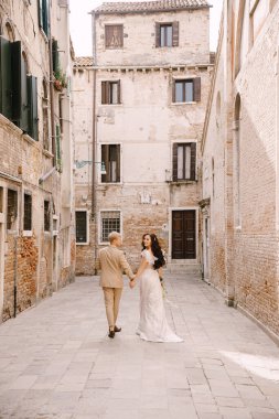 Italy wedding in Venice. The bride and groom walk along the deserted streets of the city. Newlyweds are walking in a dead end alley on the background of brick buildings. clipart