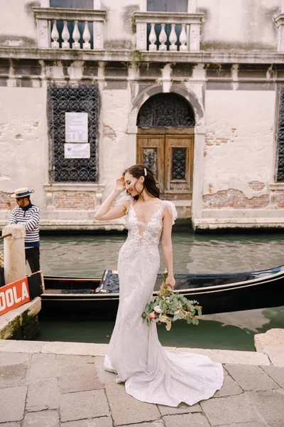 Venice wedding in Italy. A bride in a white dress, with a train, with a bouquet of white and red roses in her hands, stands on the pier near the moored gondola in a narrow Venetian canal. — Stock Photo, Image